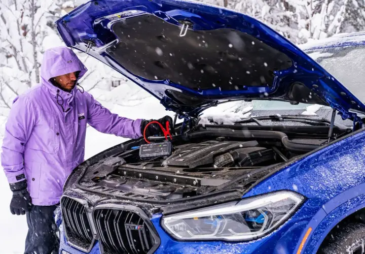Mężczyzna w fioletowej kurtce podłącza booster do akumulatora niebieskiego BMW. To rozwiązanie, co może pobierać prąd w samochodzie na postoju, gdy jest zimno.