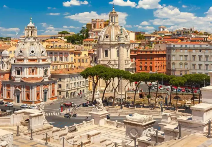 Vista panoramica di Roma con l'Altare della Patria in primo piano, cupole imponenti e palazzi storici sotto un cielo azzurro con nuvole bianche.