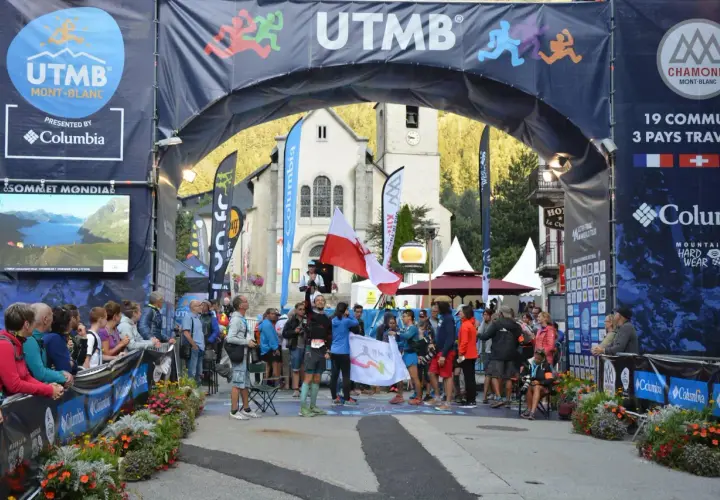 Uczestnicy Ultra-Trail du Mont-Blanc świętują pod łukiem startowym. W tle kościół i tłum widzów.