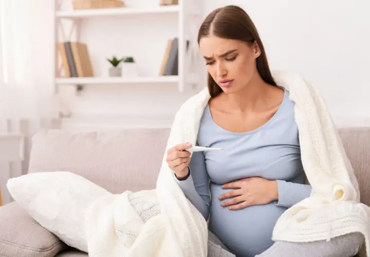 Donna incinta preoccupata guarda il termometro, avvolta in una coperta. La febbre in gravidanza è un sintomo da non sottovalutare.