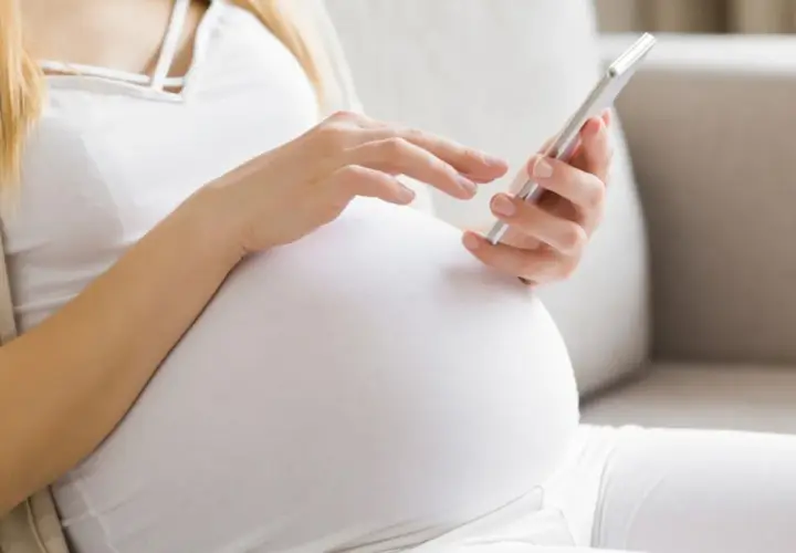 Kobieta w 24. tygodniu ciąży (l4 w ciąży) przegląda coś na telefonie, siedząc na kanapie.