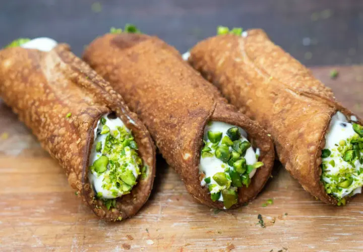 Trzy chrupiące włoskie desery cannoli, wypełnione kremem i posypane pistacjami, kuszą na drewnianej desce.
