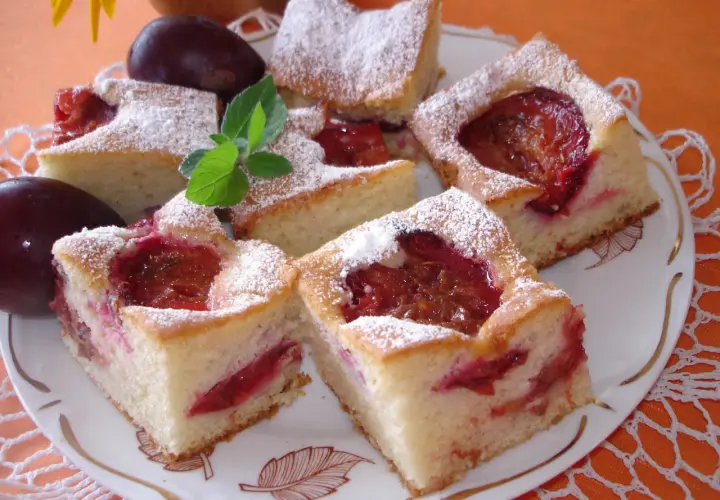 Pyszne ciasto ze śliwkami na oleju i jogurcie, posypane cukrem pudrem, prezentuje się apetycznie na talerzu.