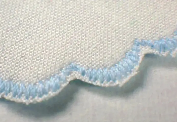 Close-up of a white fabric with a scalloped edge finished with light blue stitching. This decorative edge is called a picot.