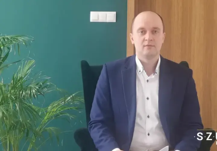 Mężczyzna w garniturze siedzi w fotelu obok rośliny, na tle zielonej ściany i drewnianych drzwi.