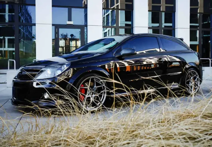 Czarny Opel Astra GTC z chromowanymi felgami i czerwonymi zaciskami hamulcowymi, zaparkowany przed nowoczesnym budynkiem, otoczony suchą trawą.
