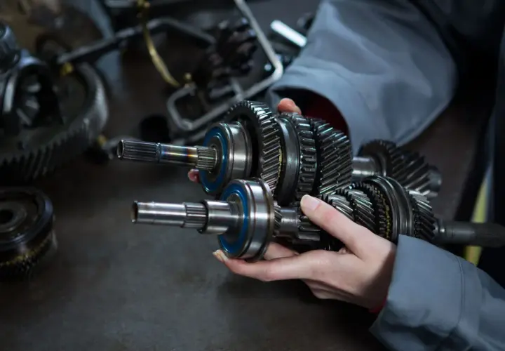 Dłonie trzymające wałek z zębatkami i łożyskami, na tle innych części mechanicznych.