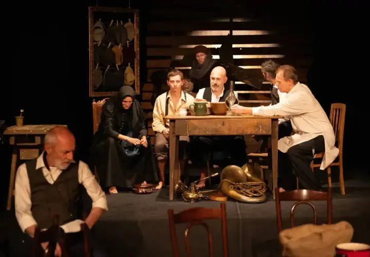 Scena teatralna, grupa ludzi przy stole, atmosfera napięcia i oczekiwania. Ten dramat porusza trudne tematy.