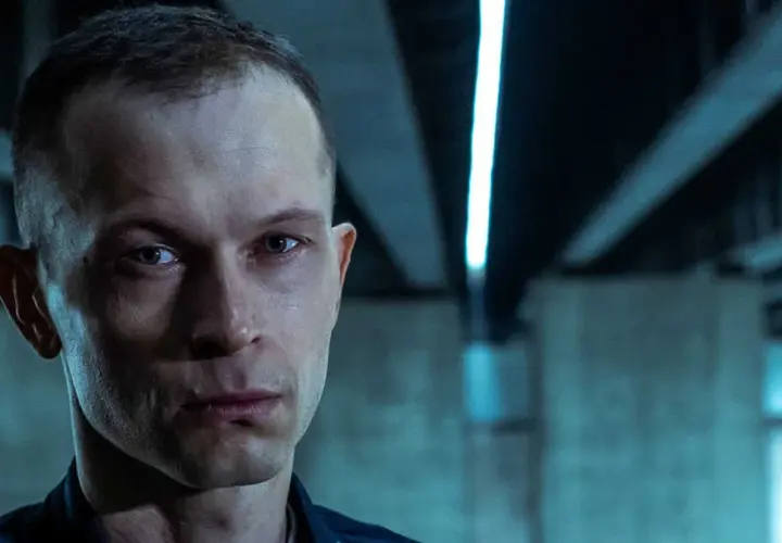 Zbliżenie na twarz mężczyzny z filmu "Tomasz Komenda". Jego spojrzenie jest intensywne, a tło sugeruje mroczne otoczenie.