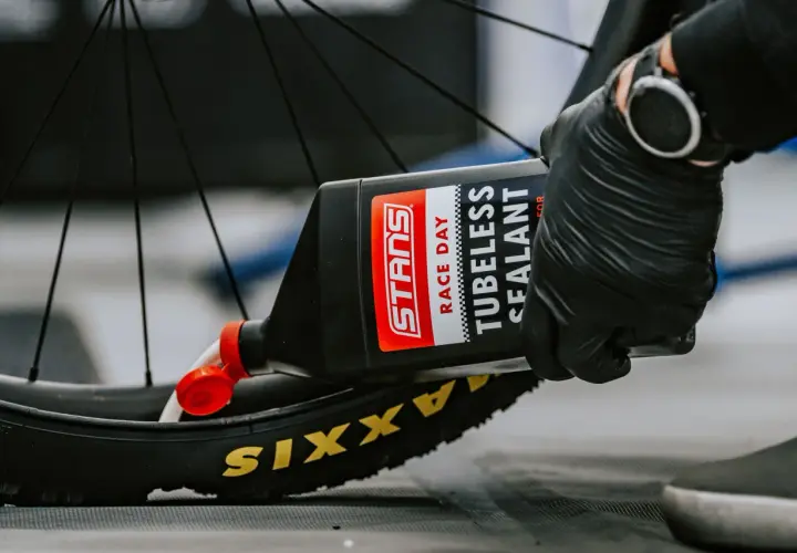 Gloved hands pour Stans Race Day tubeless sealant into a Maxxis tire, demonstrating how to add sealant to tubeless tires for a puncture-free ride.