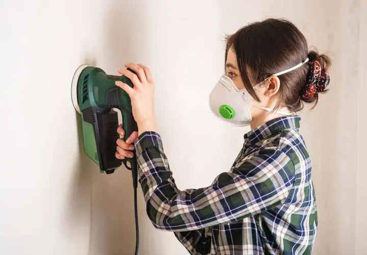 Mujer con mascarilla lijando yeso en pared con lijadora eléctrica.