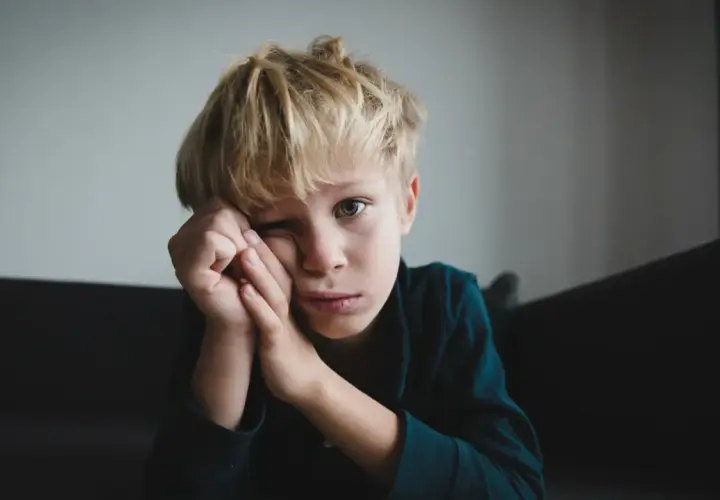 Chłopiec z blond włosami, zamyślony, z rękami przy twarzy. Jego wyraz twarzy może sugerować niektóre objawy aspergera, jak trudności w wyrażaniu emocji.