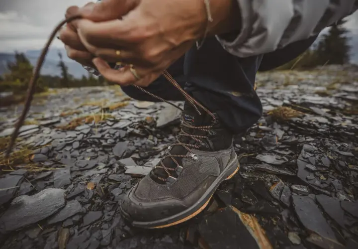 Ręce zawiązują sznurówki wysokich butów trekkingowych na kamienistym, wilgotnym podłożu.