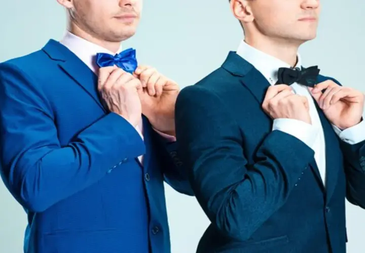 Dwóch mężczyzn w eleganckich garniturach, jeden w niebieskim, drugi w granatowym, poprawia muszki. Jaka mucha do granatowego garnituru?