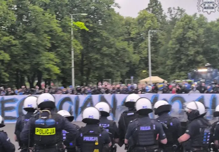 Policja w pełnym rynsztunku stoi przed grupą pseudokibiców, którzy trzymają transparent.