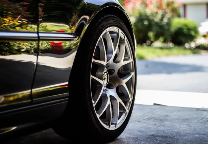 Detalle de un coche negro con llantas plateadas, listo para cambiar ruedas delanteras. Reflejos de flores y vegetación en la carrocería.