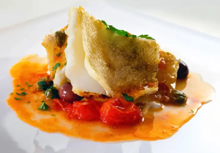 Delicioso bacalao encebollado con tomates, aceitunas y alcaparras en una salsa naranja vibrante.