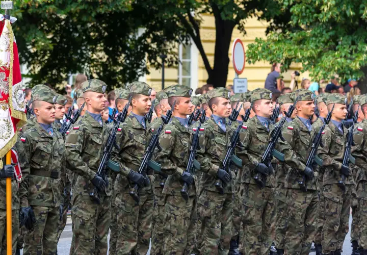 Żołnierze w mundurach i beretach stoją w zwartym szyku, trzymając broń. W tle widać drzewa i budynki, a także flagę. To widok stanowisk wojskowych podczas parady.