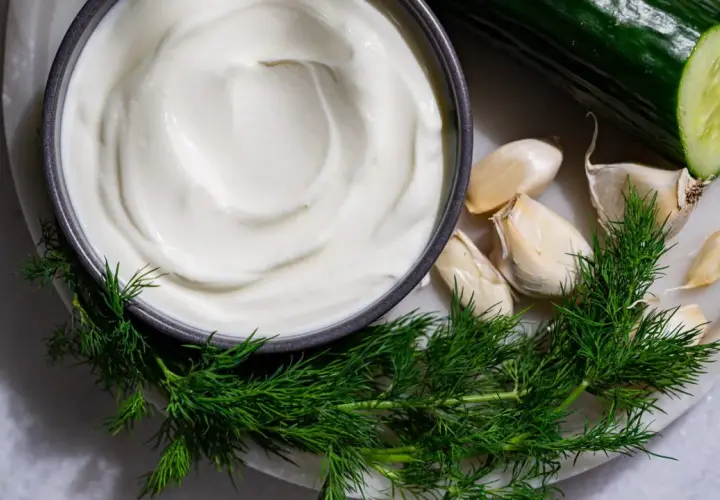 Crema blanca cremosa en un tazón, rodeada de eneldo fresco, ajos y un pepino cortado. Ingredientes para una deliciosa salsa tzatziki.