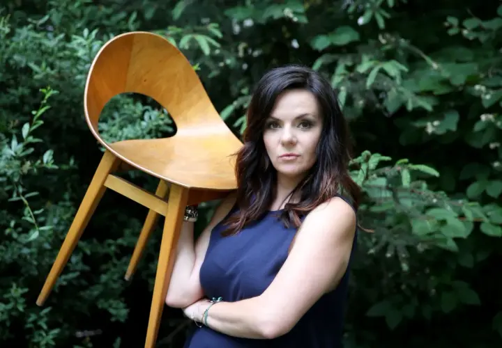 Krystyna Łuczak-Surówka, w wieku, z dumą prezentuje designerskie krzesło na tle zieleni.