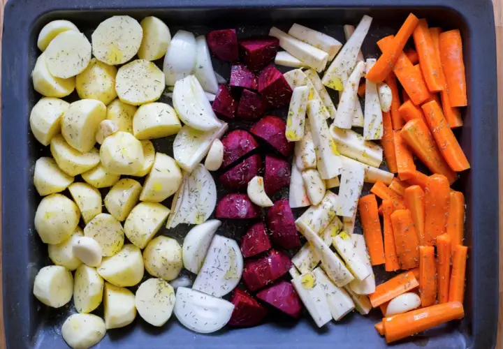 En ugnsplåt med förberedda rotfrukter i ugn: potatis, lök, rödbetor, palsternacka och morötter, kryddade och oljade.