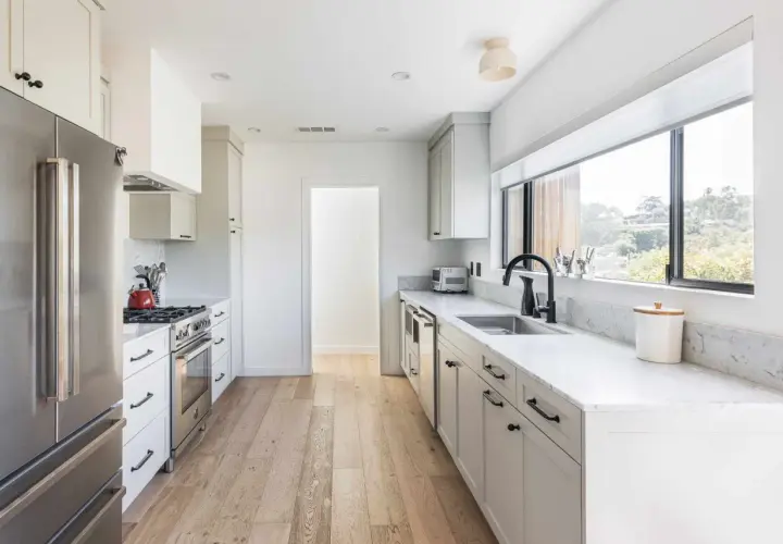 A bright, modern galley kitchen with white cabinets, stainless steel appliances, and a large window overlooking greenery.