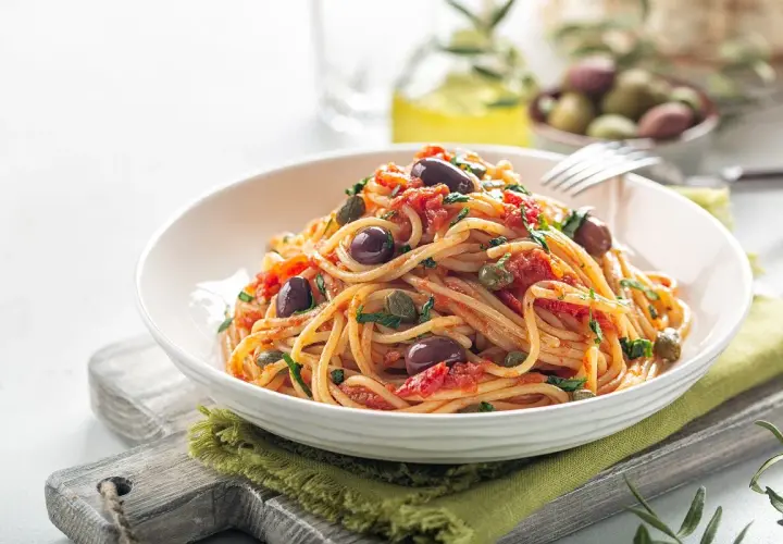 Makaron spaghetti z pomidorami, oliwkami i kaparami, podany w białej misce na zielonej serwetce.