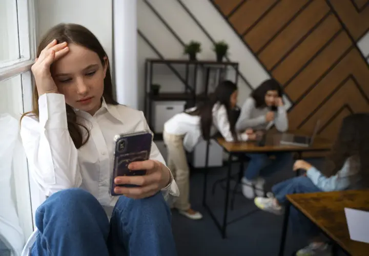 Dziewczyna z fobią społeczną siedzi sama, trzymając telefon, podczas gdy inni rozmawiają w tle.