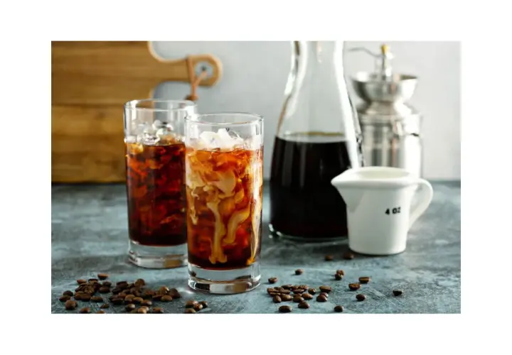 Dwa szklanki orzeźwiającego cold brew z lodem i mlekiem, obok dzbanek z kawą i młynek. Idealne na gorące dni, przygotowane z myślą o miłośnikach kawy.