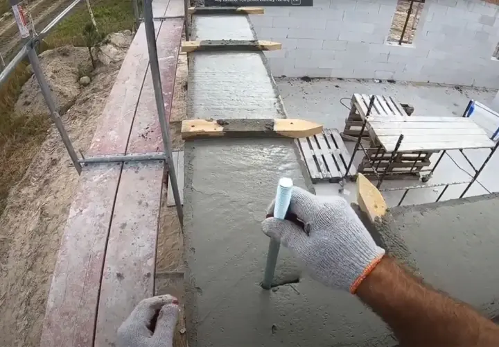 Ręka w rękawicy wkręca pręt w świeży beton. To mocowanie murłaty bez wieńca, kluczowe dla stabilności konstrukcji.