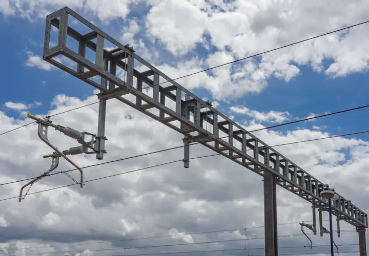 Metalowa konstrukcja sieci trakcyjnej, w tym wskaźnik W16, na tle błękitnego nieba z chmurami.