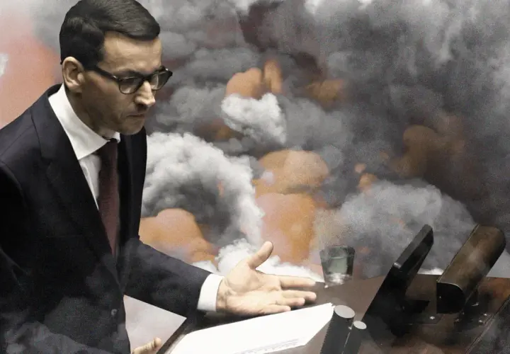 Mateusz Morawiecki w garniturze przemawia na tle dymu i pomarańczowych płomieni.