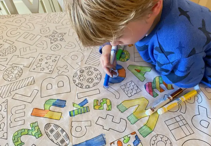 Niño rubio colorea un mantel con letras y dibujos. El **cuadrado para colorear** se llena de vida con sus rotuladores.