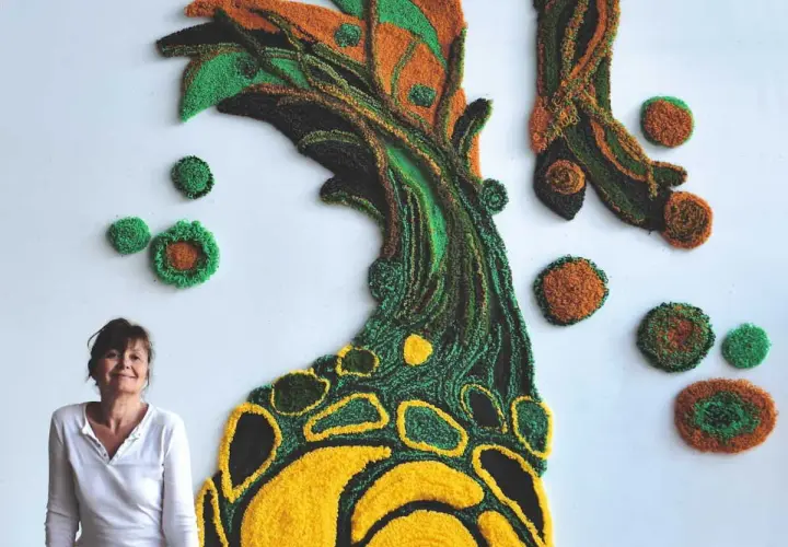 Une designer textile pose devant sa création murale en relief, un arbre aux couleurs vives.