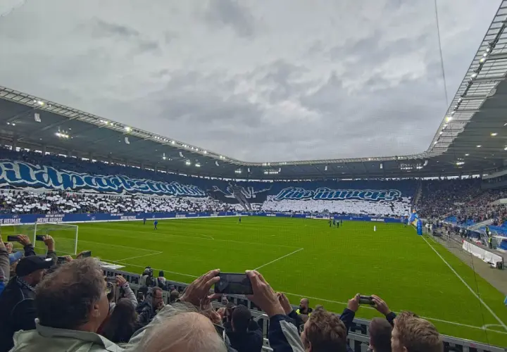 Trybuny stadionu wypełnione kibicami Karlsruher SC. Fani tworzą ogromną oprawę z napisami i sylwetką zawodnika.