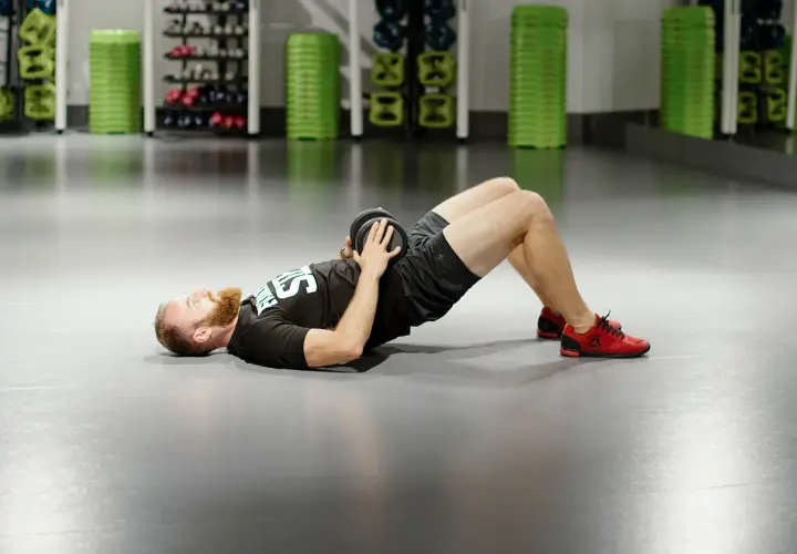 Mężczyzna z brodą wykonuje ćwiczenie glute bridge z obciążeniem na biodrach.
