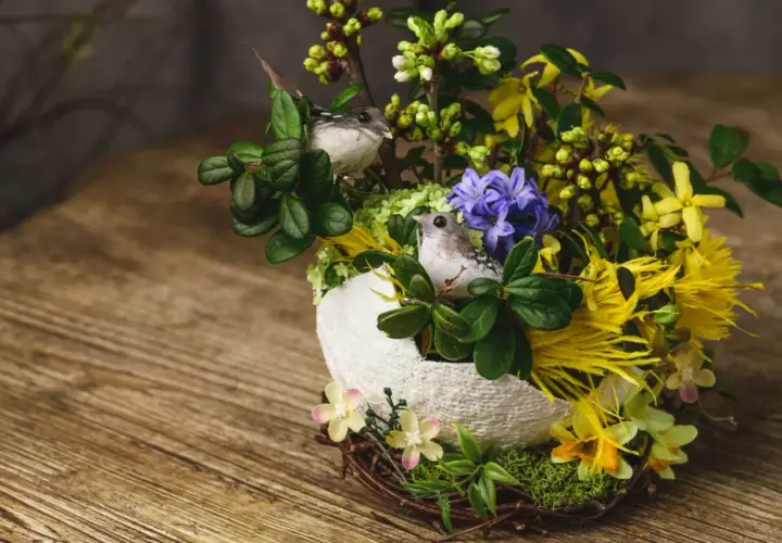 Un nid de paques composition florale de paques avec deux petits oiseaux, des fleurs jaunes et violettes, et des branches vertes sur un fond en bois.