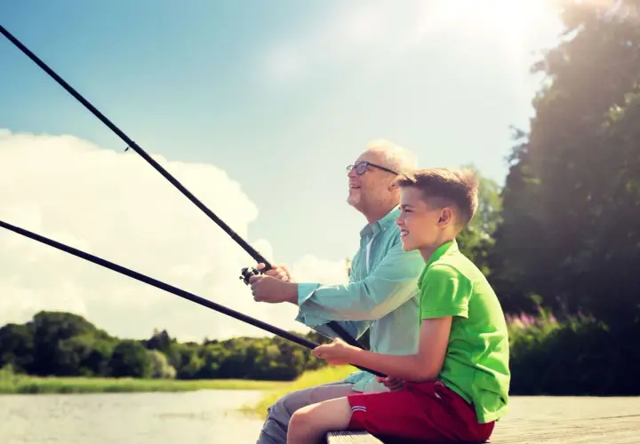 Abuelo y nieto disfrutan de un día de miños pesca, con cañas en mano y sonrisas en el rostro, bajo un cielo azul y soleado.