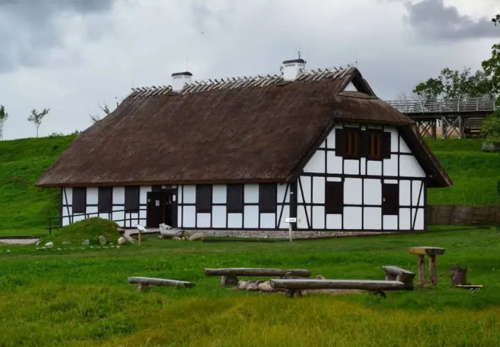 Rzucewo osada łowców fok. Dom z muru pruskiego i strzechą, otoczony zielenią, z ławkami i stołem w trawie.