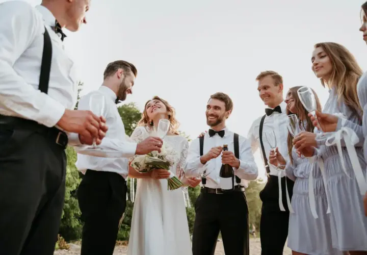 Pan młody otwiera szampana, a jego świadek i druhna wznoszą toast. Wszyscy uśmiechnięci, świętują ten wyjątkowy dzień.