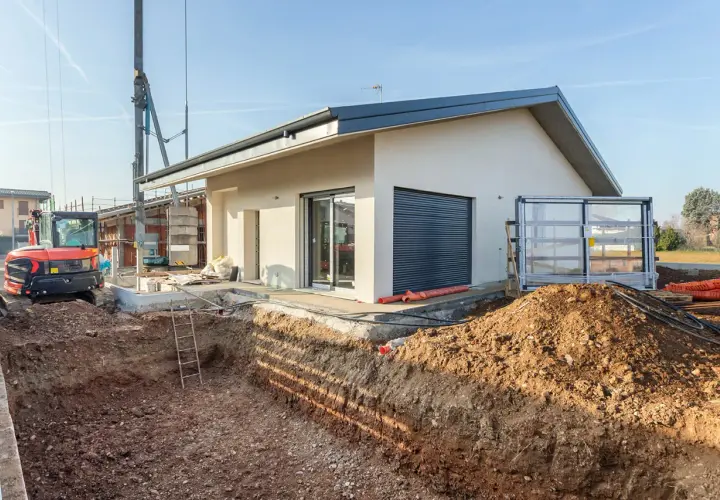 Budowa domu: wykop pod fundamenty, koparka i częściowo wykończony budynek.