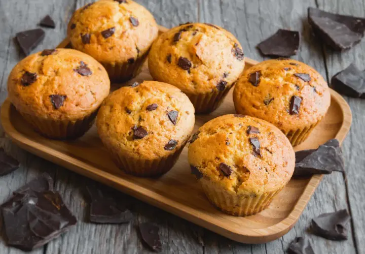 Sei muffin allo yogurt con gocce di cioccolato su un vassoio di legno, circondati da pezzi di cioccolato fondente.