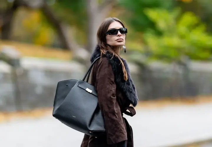 Mujer con abrigo marrón y bolso negro de Demellier, gafas de sol y pendientes llamativos.