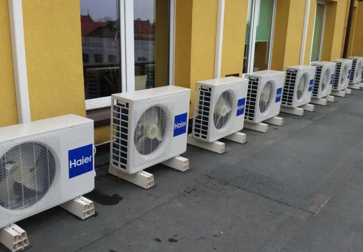 Rząd białych jednostek klimatyzacyjnych Haier na dachu, obok żółtej ściany budynku.