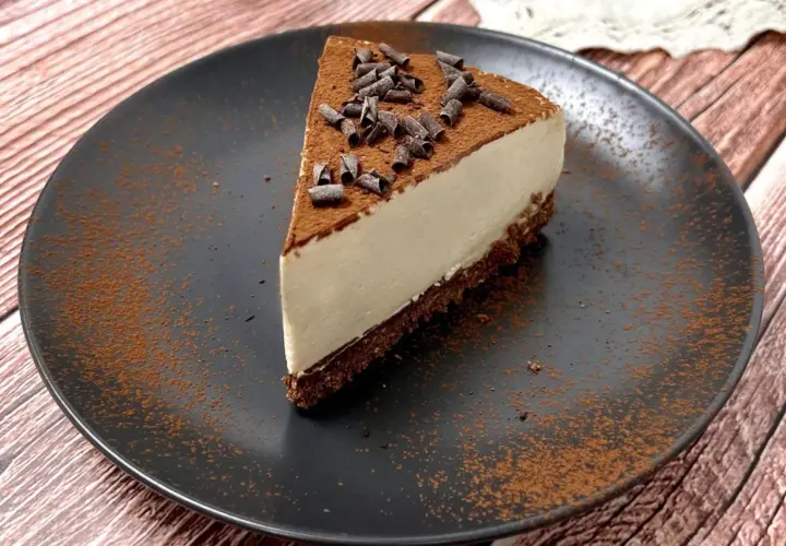 Una fetta di cheesecake al caffè, cosparsa di cacao e riccioli di cioccolato, su un piatto nero.