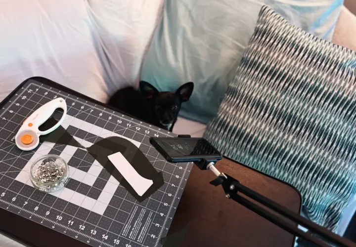 A small black dog watches as someone uses adaptive sewing tools, including a rotary cutter and pins, on a cutting mat.
