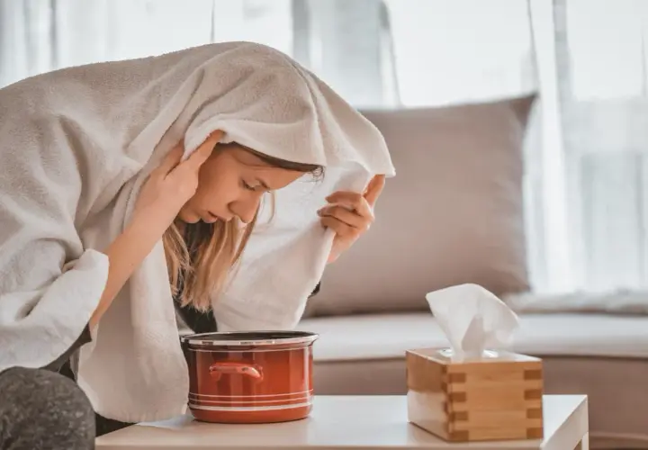 Kobieta wykonująca inhalację parową nad garnkiem z wodą, przykryta ręcznikiem.