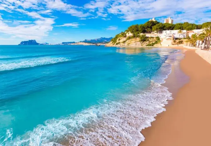 Una playa de arena dorada y aguas turquesas, con edificios blancos y vegetación en la ladera, que evoca las 10 mejores playas de Alicante.