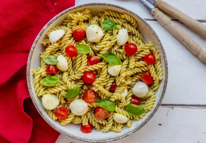 Un piatto invitante di pasta fredda al pesto, arricchito da ciliegini succosi e mozzarelline fresche, guarnito con basilico profumato.