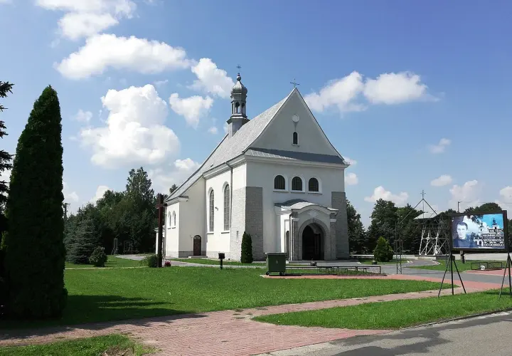 Kościół w Dulczy Wielkiej, z wieżą i krzyżem, na tle błękitnego nieba z chmurami. Obok tablica z wizerunkiem młodego mężczyzny.